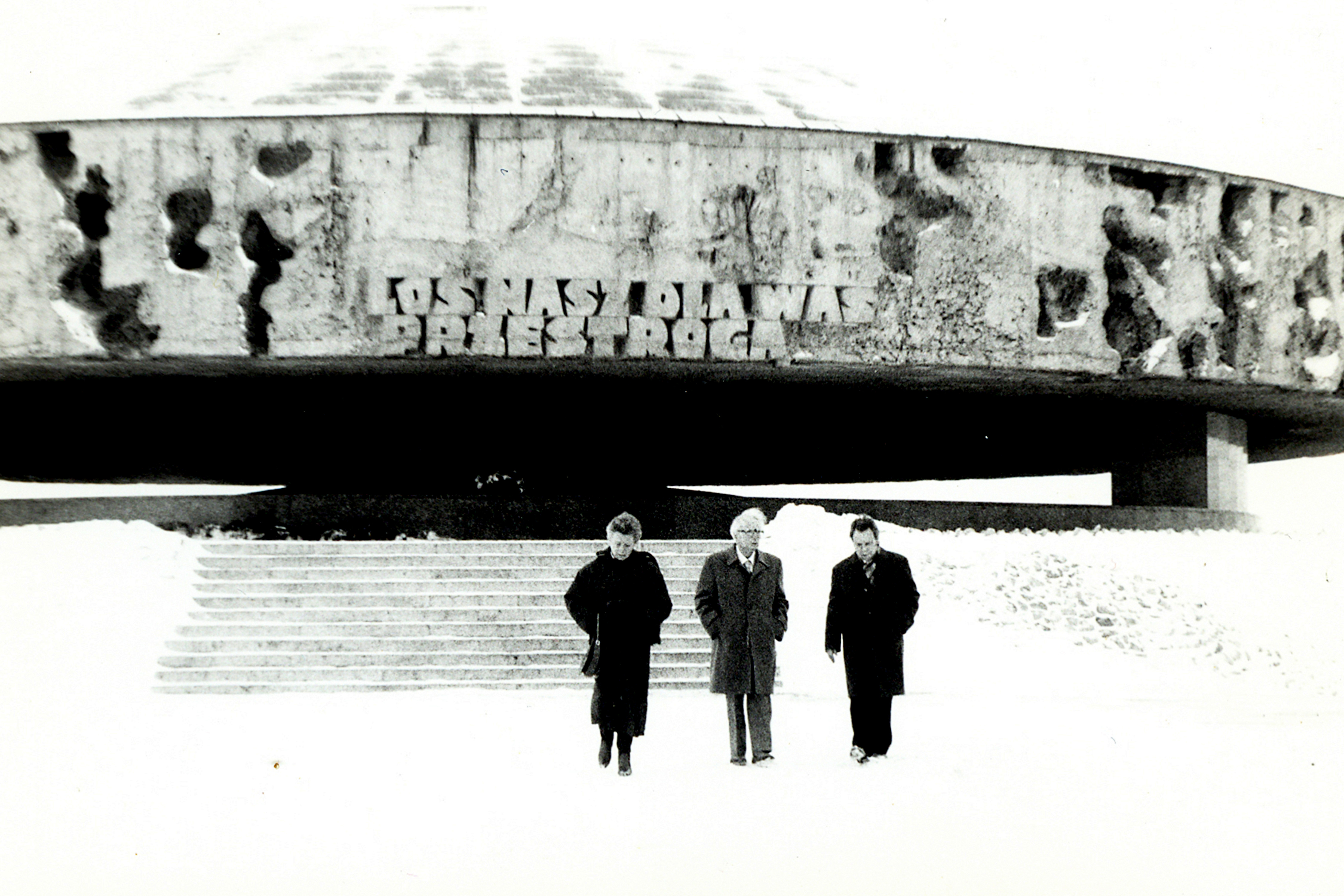 Czarno białe zdjęcie przedstawia tr&oacute;jkę os&oacute;b stojących przed pomnikiem.
